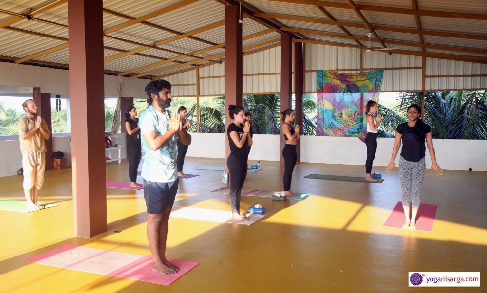 Yoga students in class with hands in anjali mudra (prayer) as the yoga teacher stands at the front of the class in samastitih (mountain pose) during yoga teacher training in Goa, India