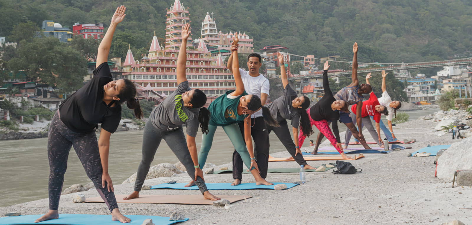 Yoga students in trikonasana (triangle pose) with a teacher adjusting during asana class by the river in Rishikesh, India.